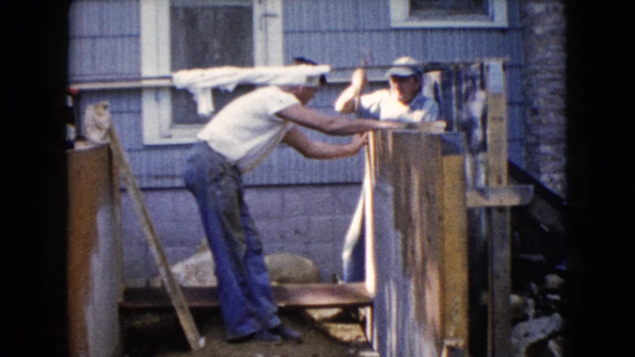 In 1960 Wauconda, Illinois home-movie footage, two men work together on a wooden structure near a house wall and window.