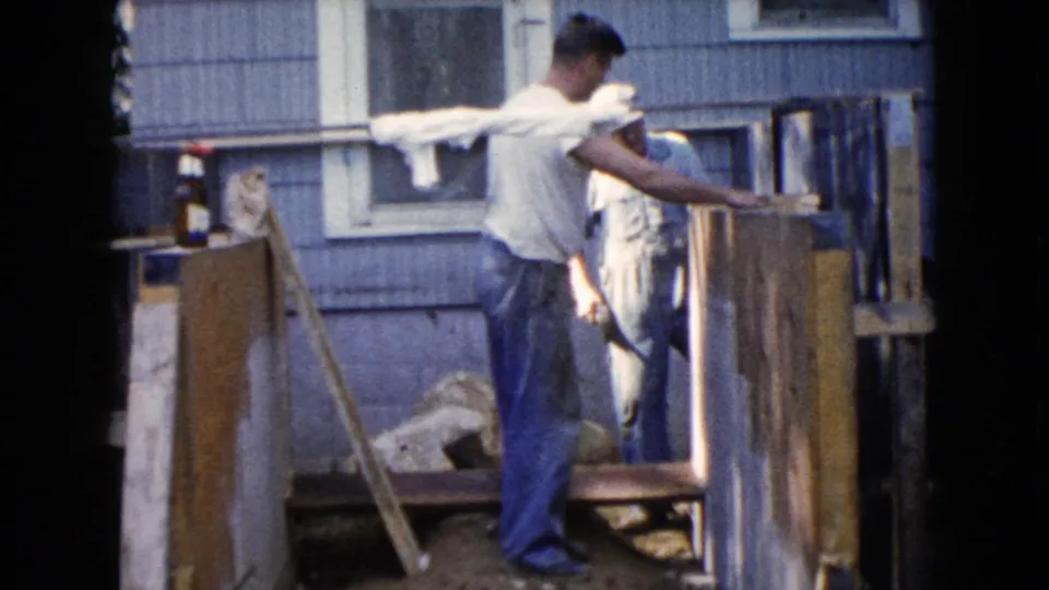 The 8mm frame shows a man beside the backyard structure, with another worker partly obscured near the boards and house wall.