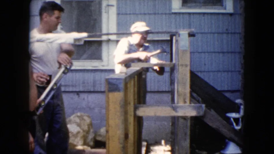A 1960 Wauconda backyard view shows two men beside a partly built wooden frame, with house siding and an exterior stairway behind them.