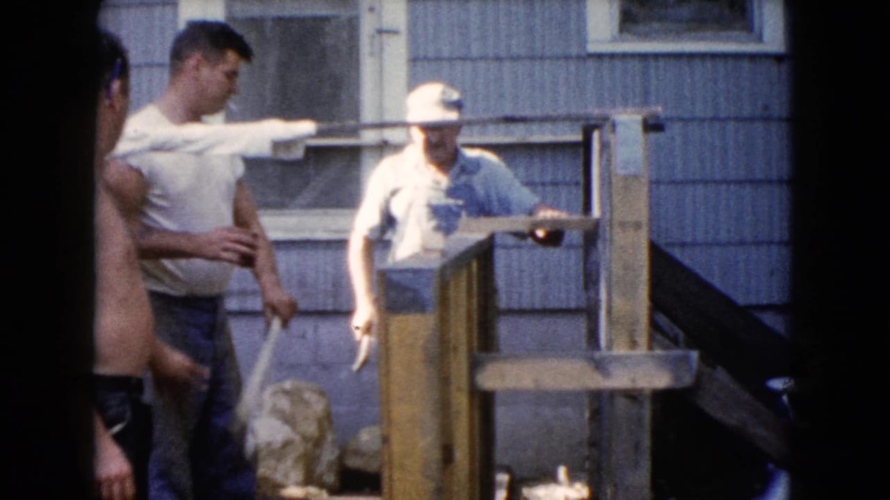 In 1960 Wauconda home-movie footage, two men stand by a wooden fence or railing outside a sided house as another shirtless man enters frame.