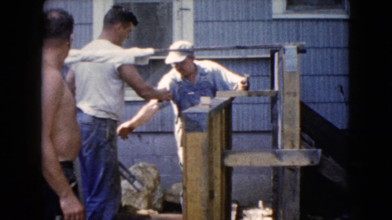 A shirtless man stands in the foreground as two men work near a wooden fence.