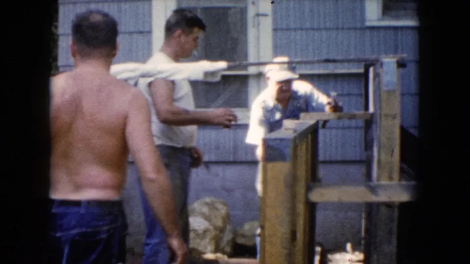 The home-movie frame shows a shirtless man from behind and two men near a wooden fence or railing against blue house siding.