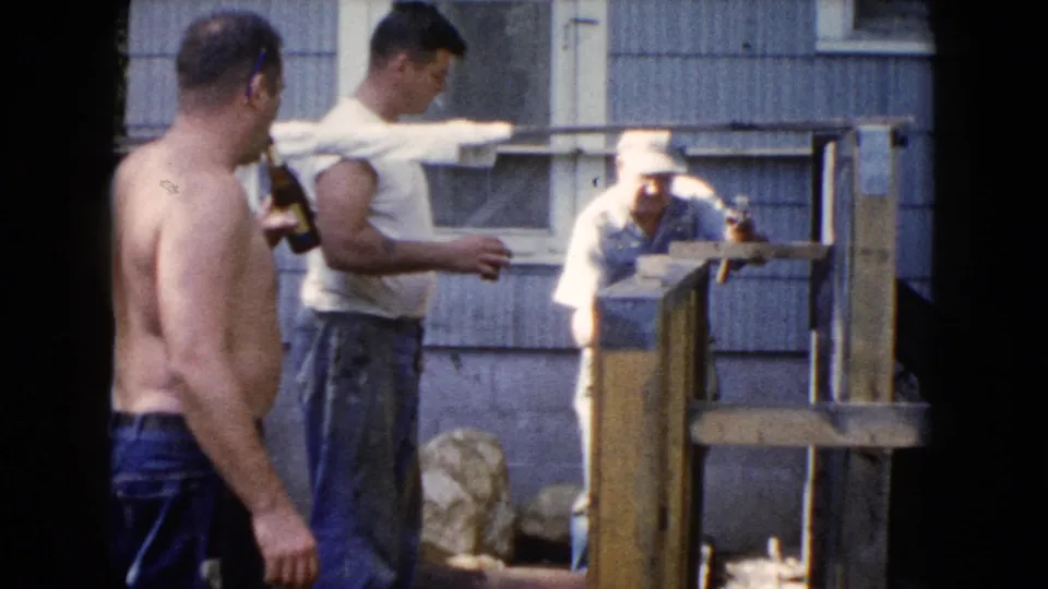 A shirtless man holding a bottle stands in the foreground while two men remain beside a wooden fence or railing outside the house.