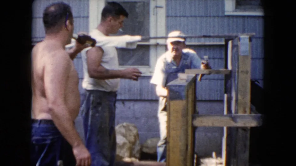 In Wauconda, Illinois home-movie footage from 1960, three adult men gather outside a house beside a partly built wooden structure.