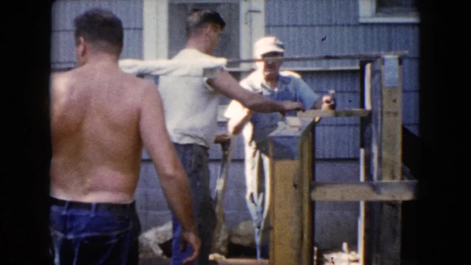 The home-movie frame shows a shirtless man approaching two men near a wood structure in a residential Wauconda yard in 1960.