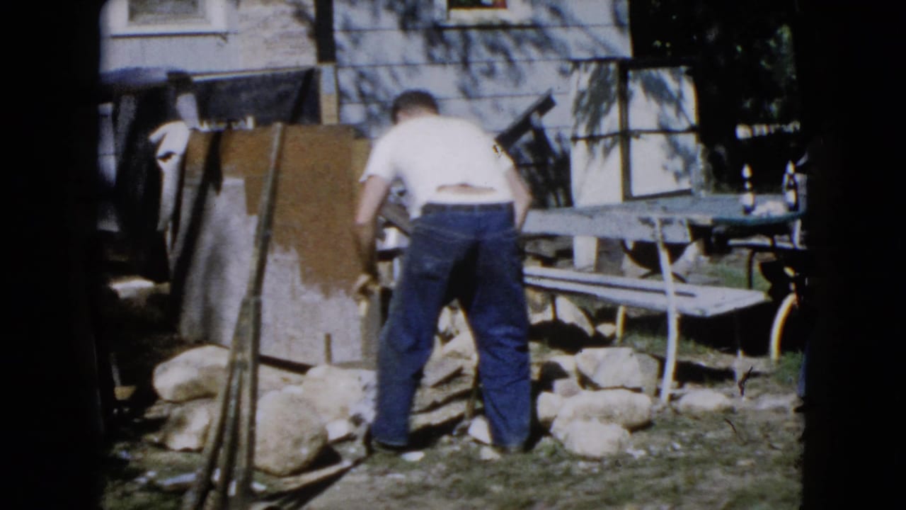 In Wauconda, Illinois home-movie footage from 1960, a man bends over large stones near a picnic table and house exterior.