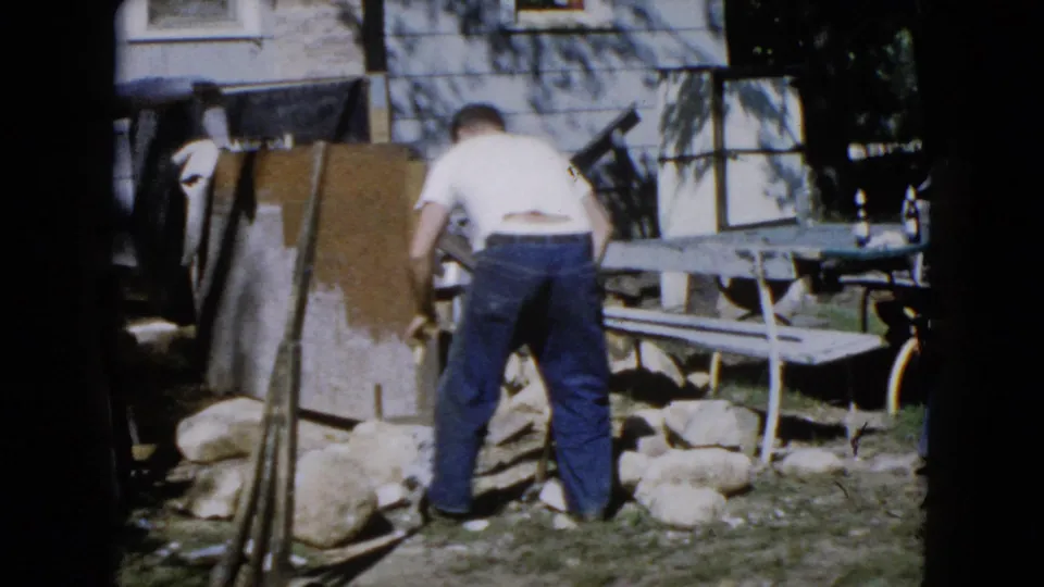 In Wauconda, Illinois home-movie footage from 1960, a man bends over large stones near a picnic table and house exterior.