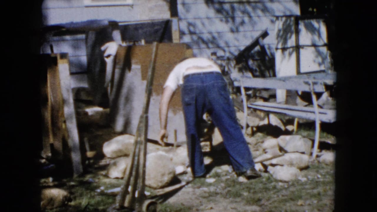 A man in a white T-shirt and jeans works near a cluster of stones in a Wauconda backyard, with boards and a picnic table behind him.