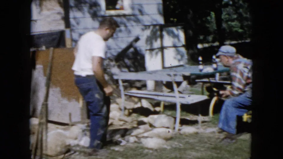 A standing man pauses near the stone work while an older man in a plaid shirt and cap sits beside the picnic table in the 1960 yard scene.