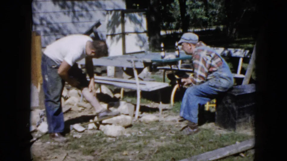 The younger man bends by the stones while the older man sits nearby in a cap and plaid shirt, framed by a picnic table and house exterior.