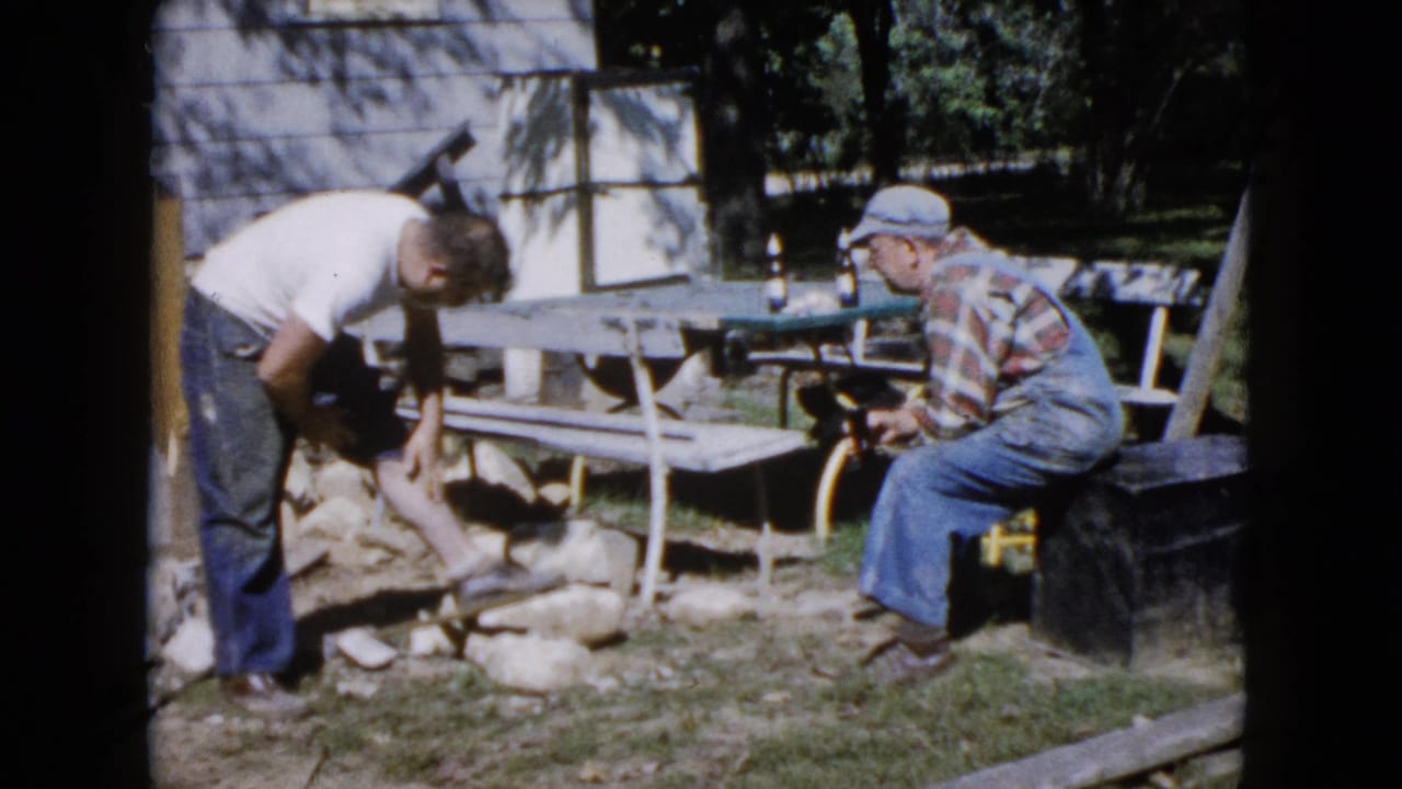 Two men outdoors near a picnic table, with one bending over masonry materials and another seated nearby.