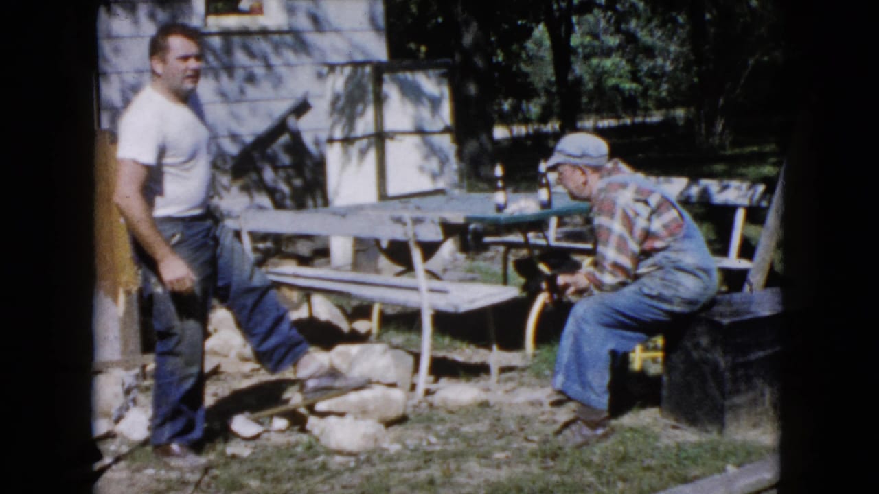 1960 Wauconda yard scene with a man in a white T-shirt standing near masonry materials and an older man seated beside a picnic table.
