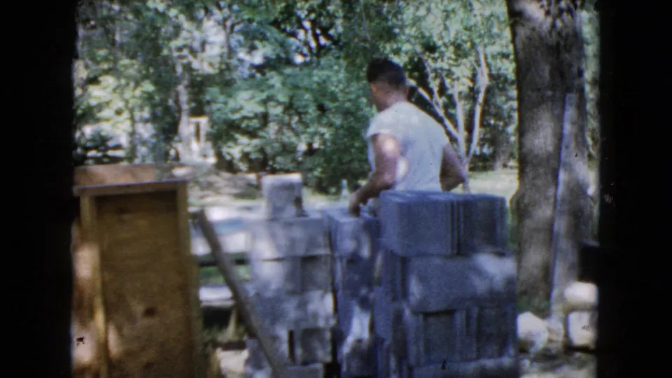 1960 Wauconda frame showing a man near stacked cinder blocks, wooden boards, and yard equipment under trees.