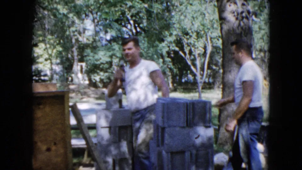 1960 Wauconda home-movie frame with two adult men beside cinder blocks, boards, and a picnic table in a leafy yard.