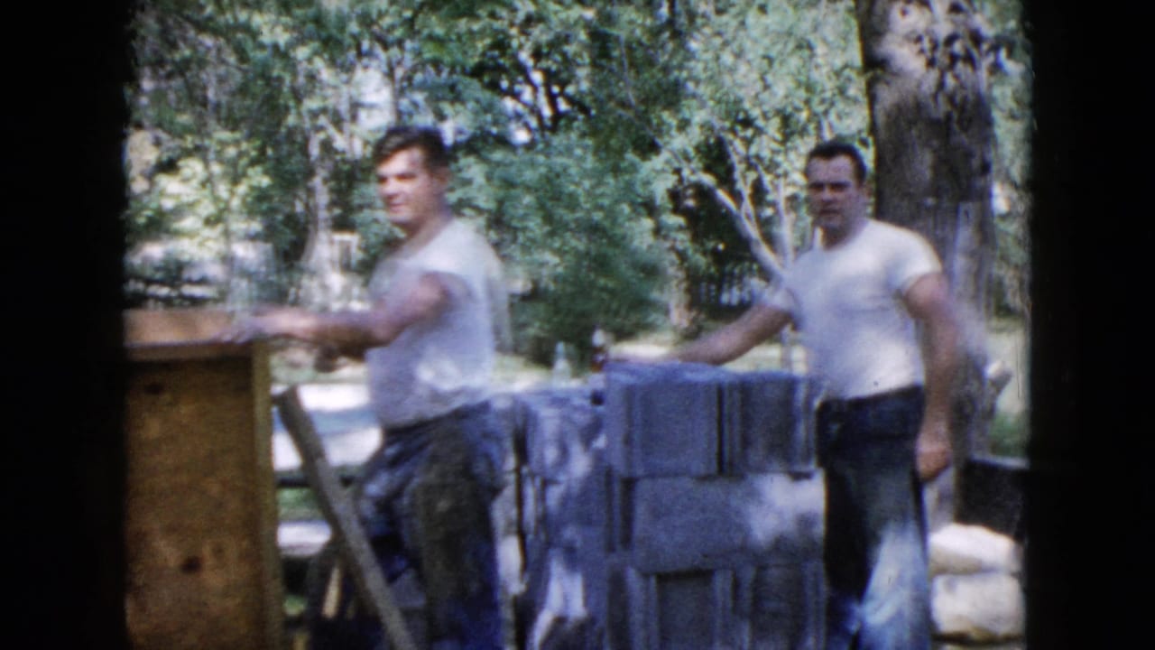 1960 Wauconda home-movie frame showing two adult men near cinder blocks and trees during outdoor work.