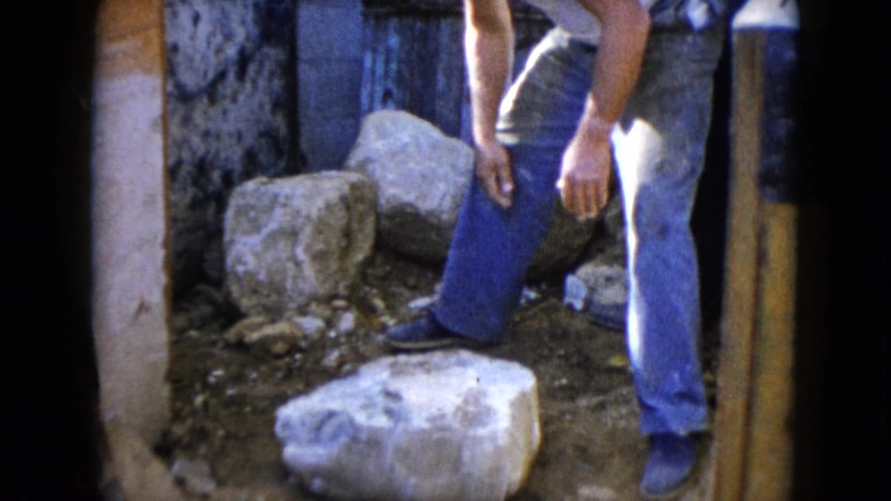 1960 Wauconda frame showing an adult man near large stones beside a rough exterior wall or structure.