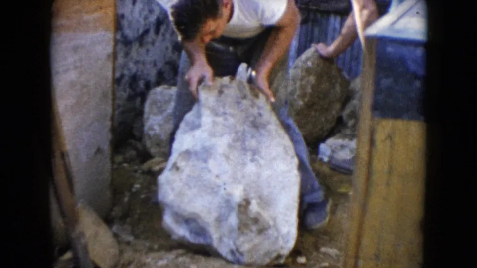 1960 Wauconda frame showing two adults handling a large stone beside a wall in an outdoor work area.