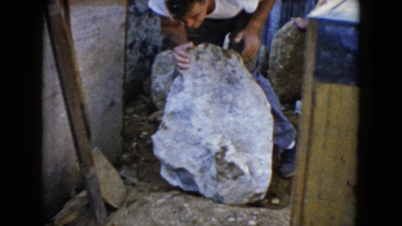 In 1960 Wauconda, Illinois home-movie footage, a man leans over a large stone near boards, dirt, and stacked materials.