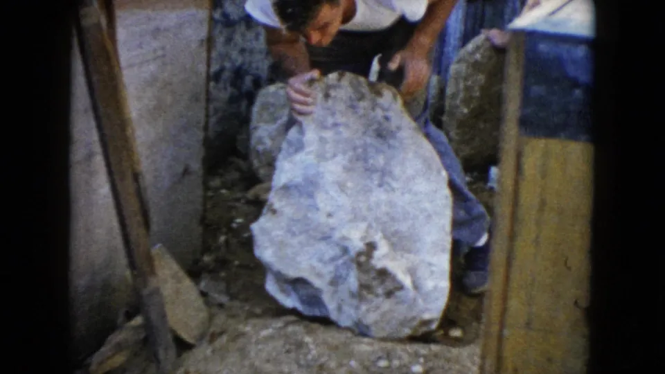 In 1960 Wauconda, Illinois home-movie footage, a man leans over a large stone near boards, dirt, and stacked materials.
