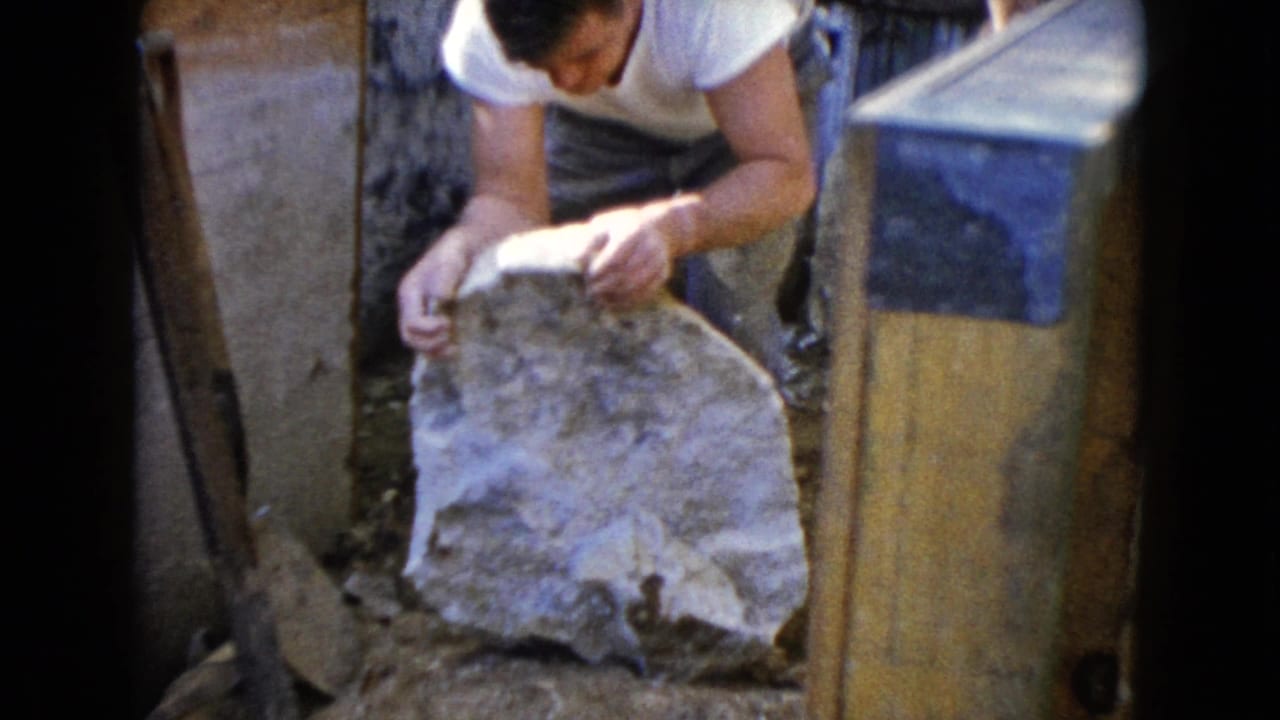 Man grips a large rock beside a wooden board and work materials.