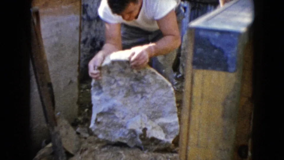 A man in a white shirt steadies a large rock in a small outdoor work area in the Wauconda, Illinois home-movie clip.