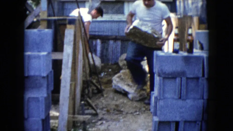 Two adult men appear near stacked cinder blocks while one carries a large stone through an outdoor work area in 1960 Wauconda footage.