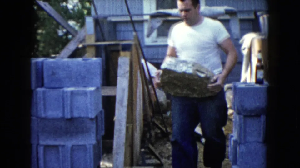 A man in a white T-shirt and blue jeans carries a large rock between cinder block stacks in the Wauconda, Illinois work scene.