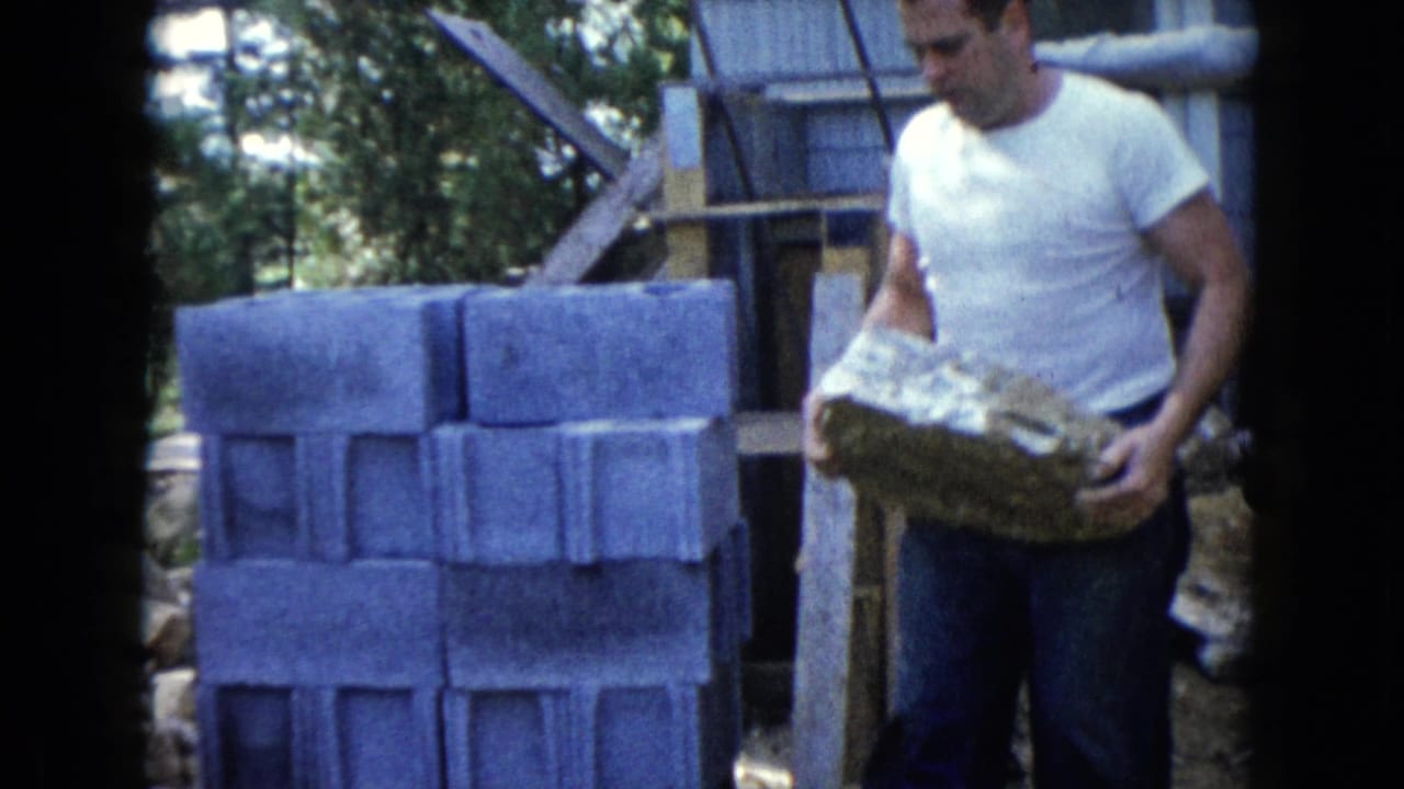 Man in a white T-shirt holds a concrete block near stacks of masonry blocks outdoors.