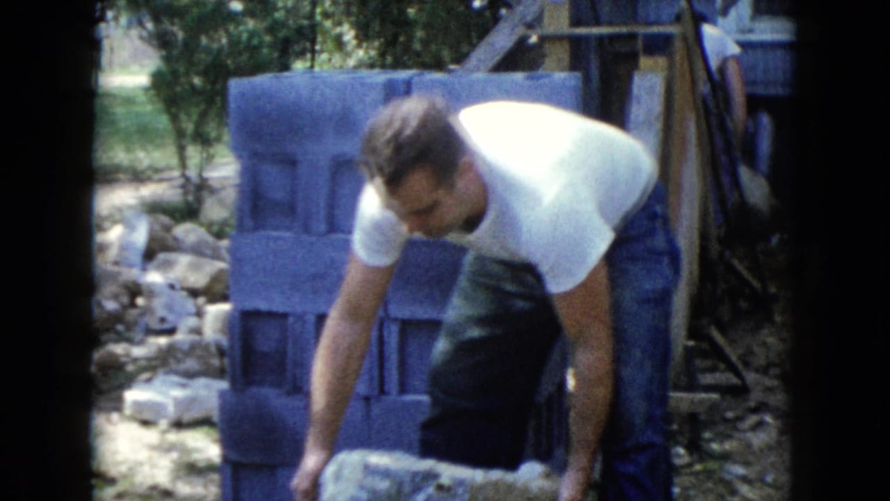 A man in a white T-shirt bends to handle a concrete block beside a stacked block wall in the Wauconda backyard scene.