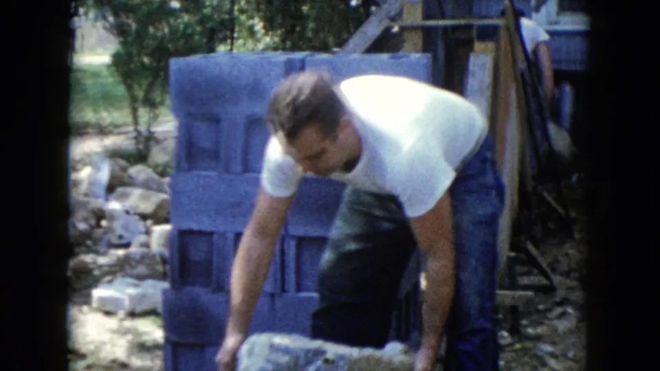 A man in a white T-shirt bends to handle a concrete block beside a stacked block wall in the Wauconda backyard scene.