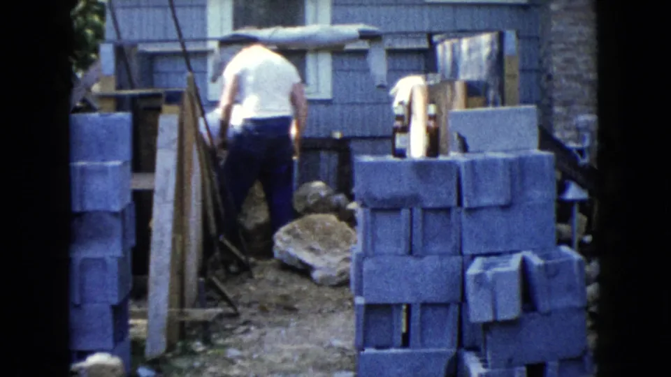 The man is seen from behind near stacked concrete blocks, rough stones, and a small building wall in the 1960 Wauconda yard footage.