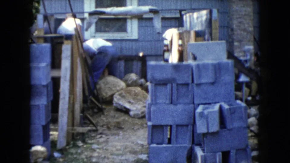 A man bends behind concrete block stacks near rough stones and a small building wall during the Wauconda backyard construction activity.