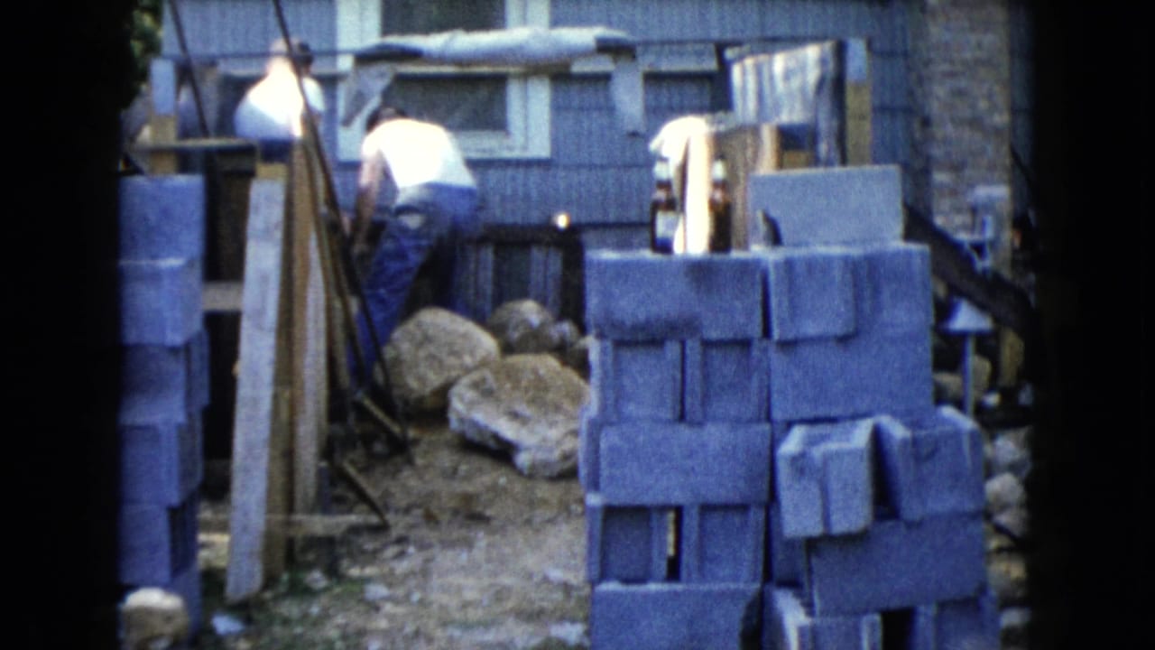 Man working near stacked cinder blocks beside a blue-shingled house.