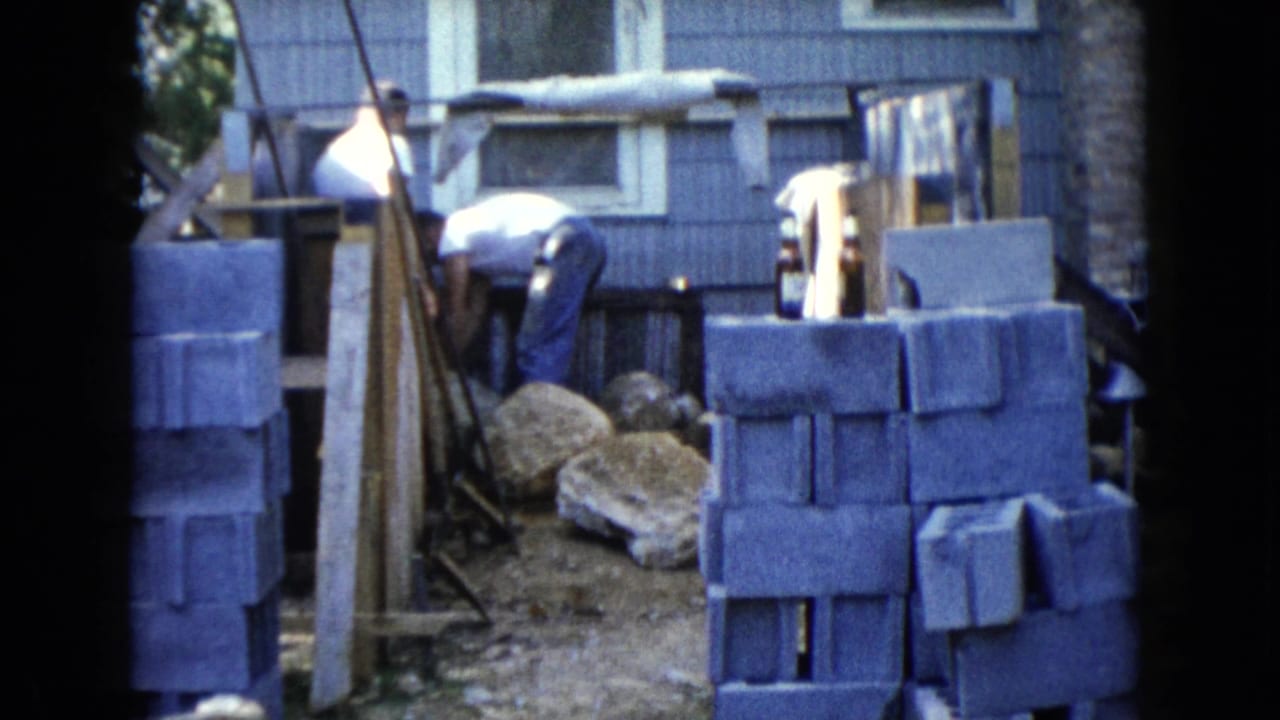 1960 Wauconda frame with a man bending near a dirt work area, stacked gray cinder blocks, boulders, and the side of a blue house.