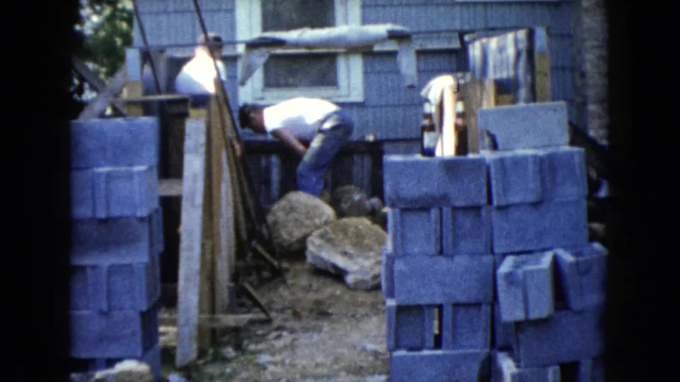 1960 Wauconda backyard frame showing a man leaning over the ground with cinder blocks, rocks, planks, and blue house siding around him.