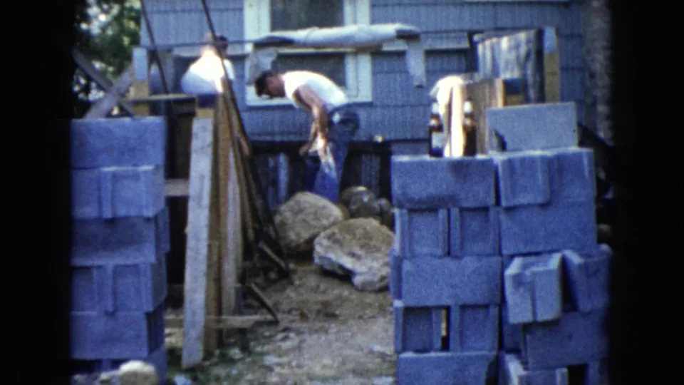 1960 Wauconda home-movie frame showing a worker bent at the waist near cinder blocks, large rocks, dirt, and blue exterior siding.
