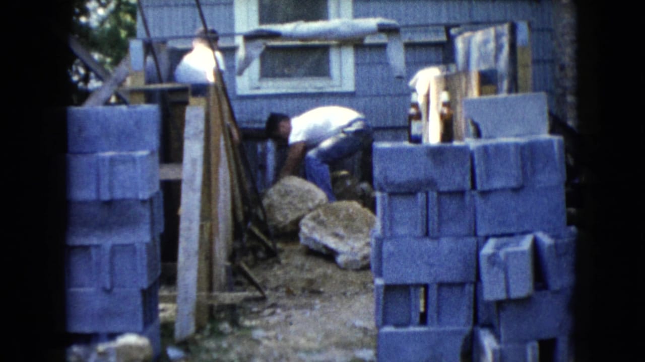 1960 Wauconda home-movie frame showing two men near concrete blocks and construction materials beside a house.