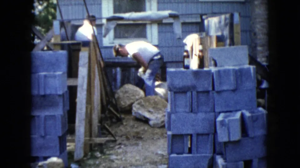 1960 Wauconda frame with a man bending over construction materials while another man stands behind block stacks.