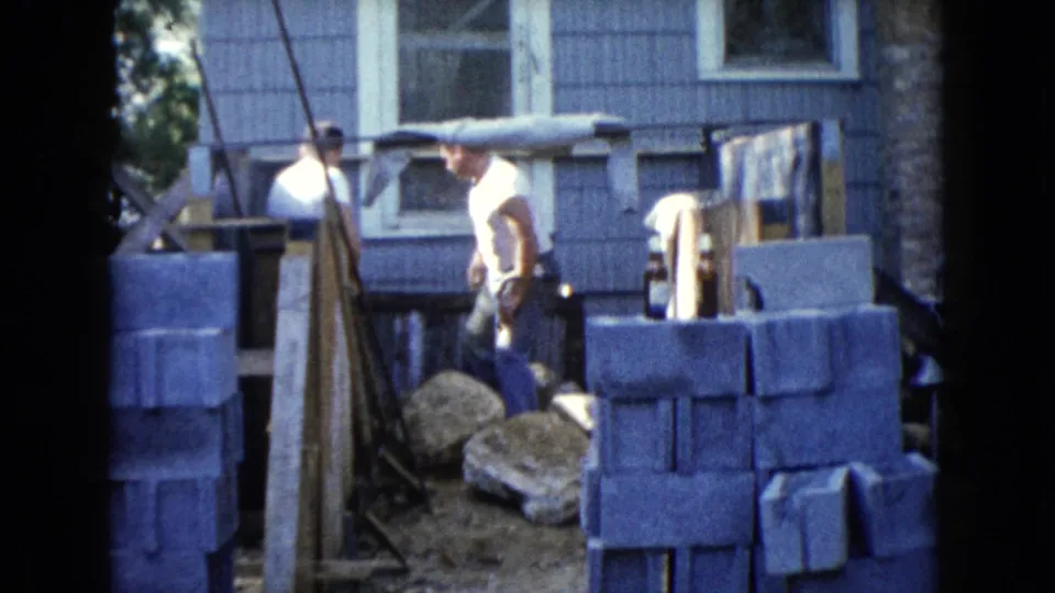 1960 Wauconda home construction frame showing a man standing among block stacks and supports beside a house exterior.
