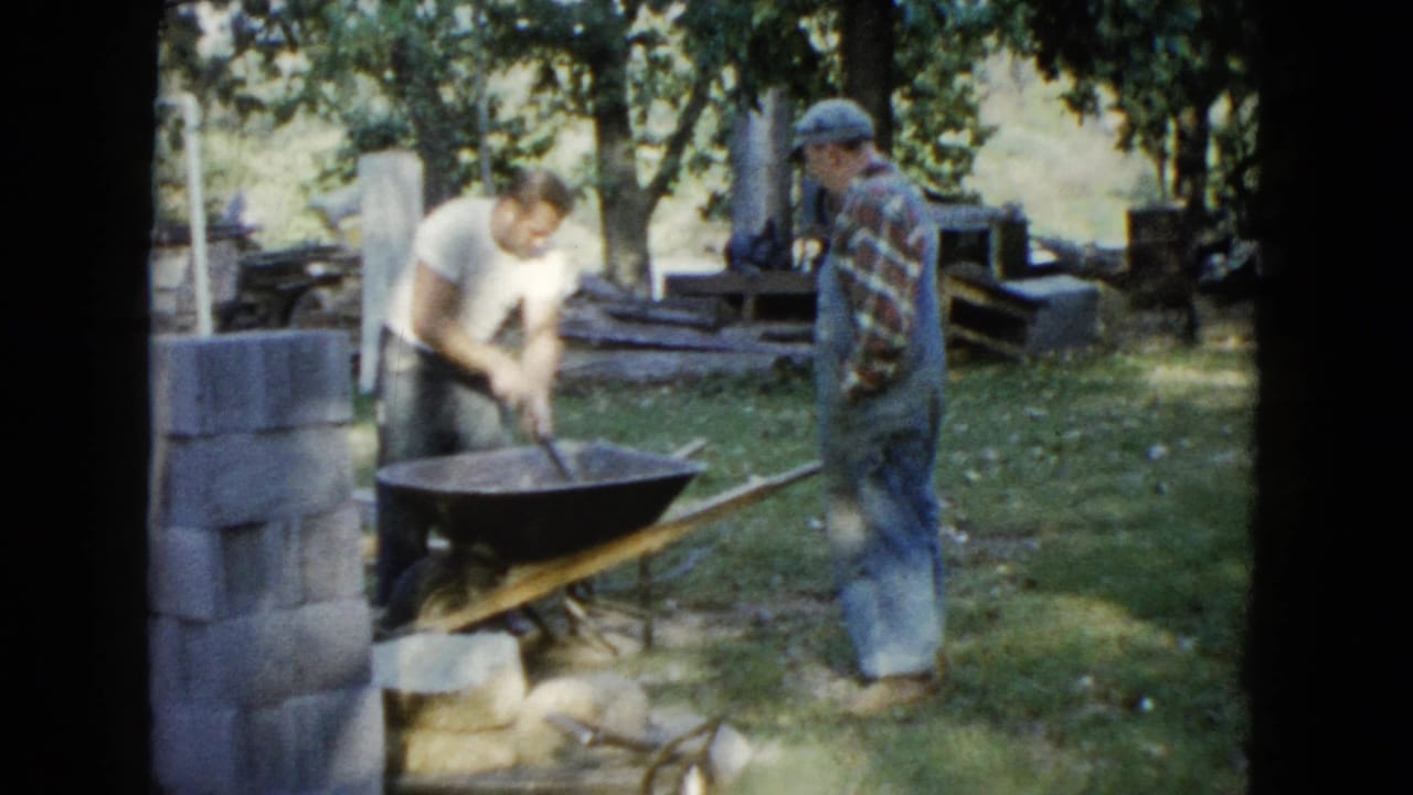 Two men beside a wheelbarrow and stacked cinder blocks in a shaded backyard.