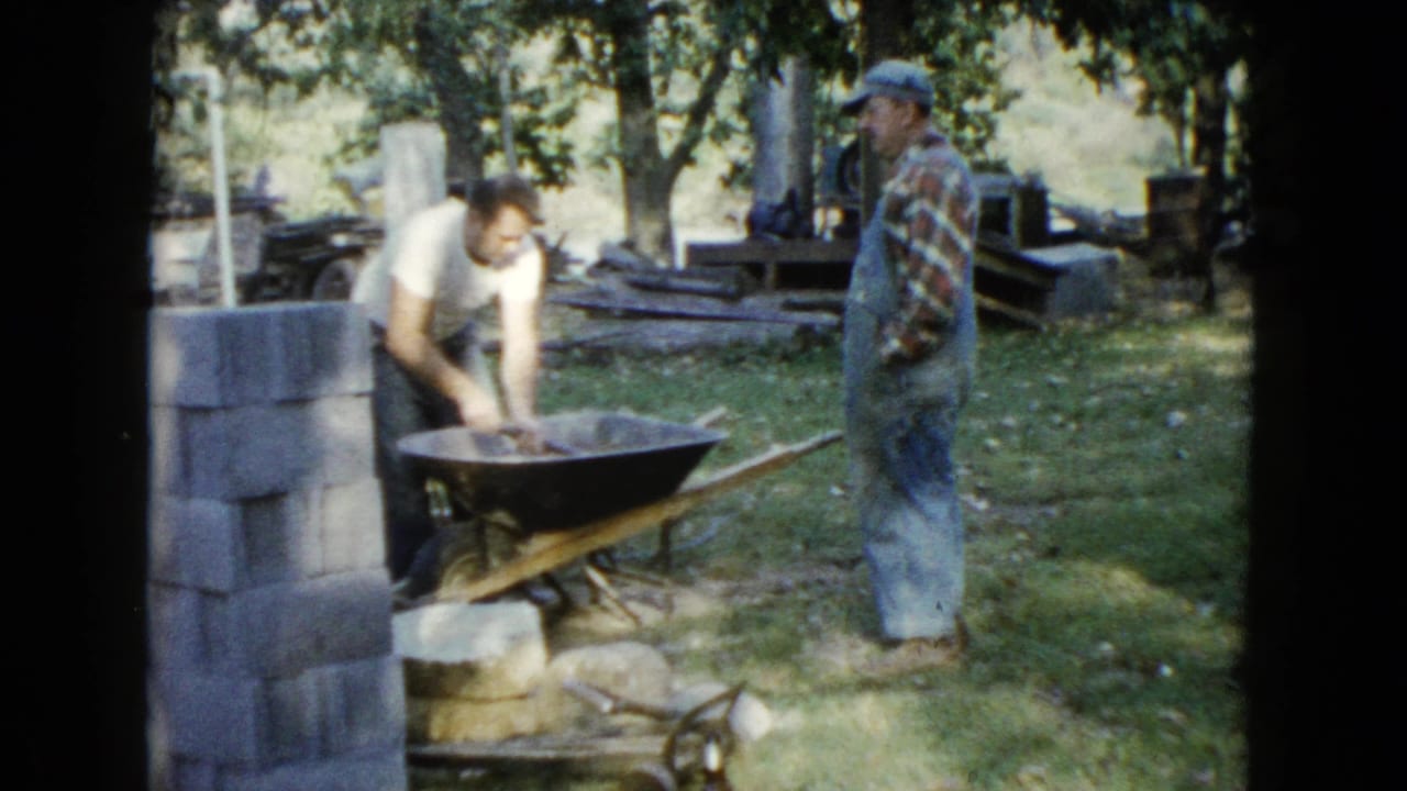 A man works in a wheelbarrow as another man stands near him in the yard.