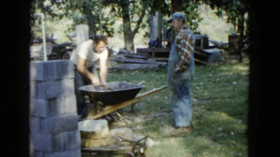 A backyard masonry scene shows a man leaning into a wheelbarrow of concrete while a second man in overalls observes nearby.