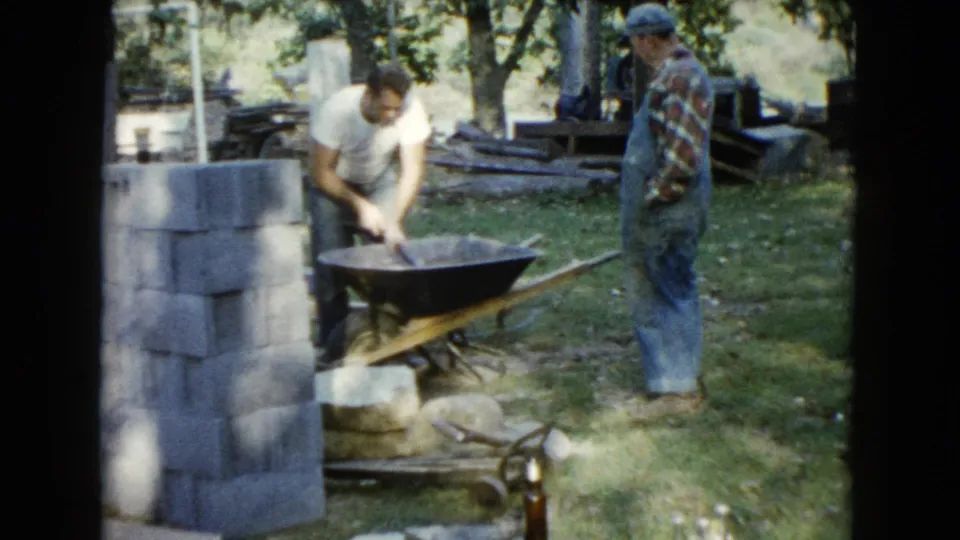 The 8mm frame shows a man working concrete in a wheelbarrow, with another man partly visible and cinder blocks stacked at left.