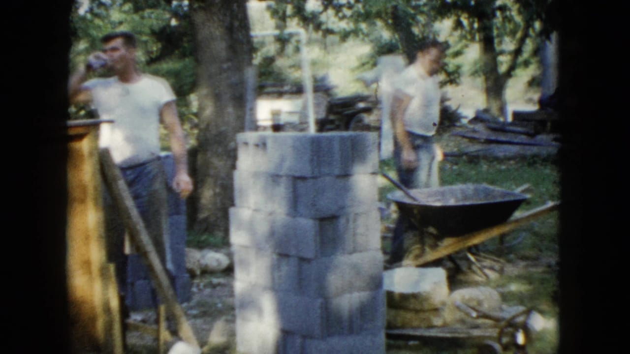Two men work near cinder blocks and a wheelbarrow in a wooded backyard construction area.