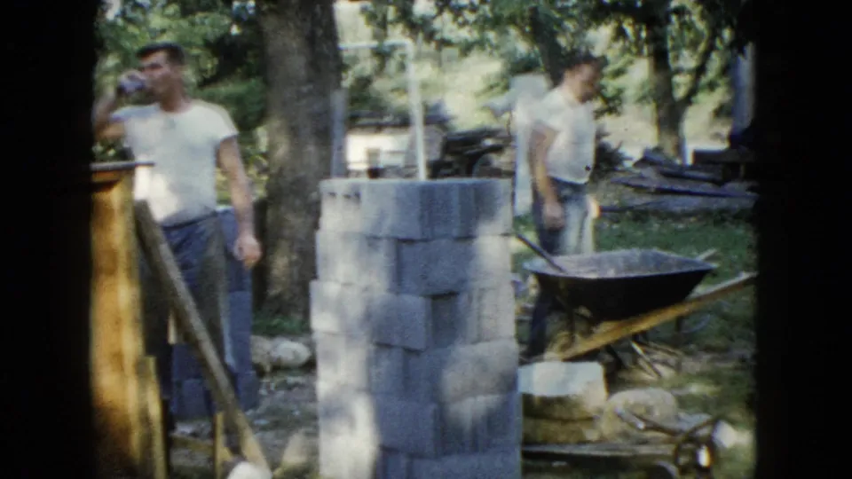 In 1960 Wauconda, Illinois footage, two adult men stand by cinder blocks, a wheelbarrow, and wood panels during backyard foundation work.