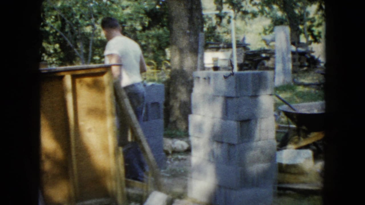A man stands beside stacked cinder blocks, wood panels, and a wheelbarrow outdoors.