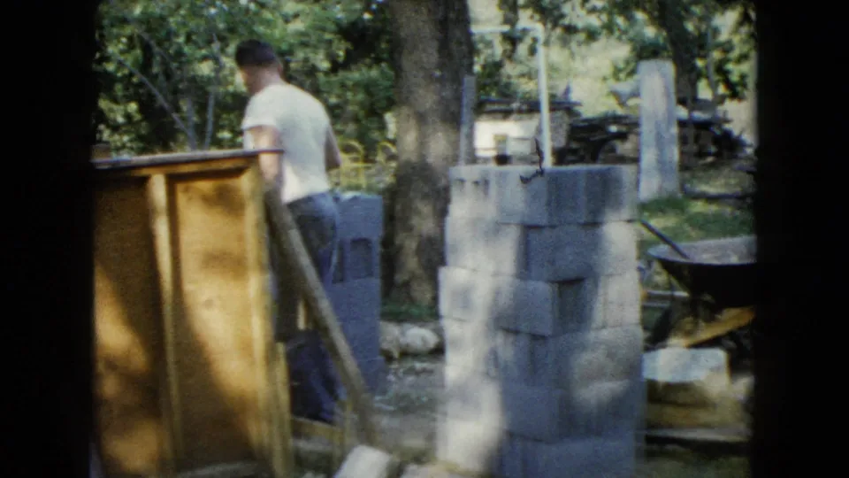 A man in a white T-shirt stands near stacked cinder blocks and wood panels in the wooded Wauconda backyard work area.