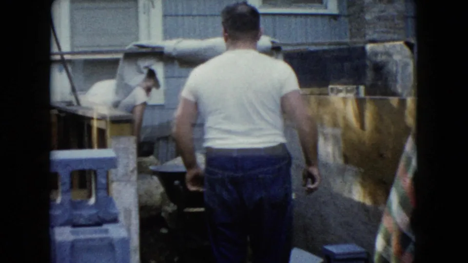 An adult man seen from behind approaches the foundation area, with cinder blocks, a wheelbarrow, and a house wall visible.