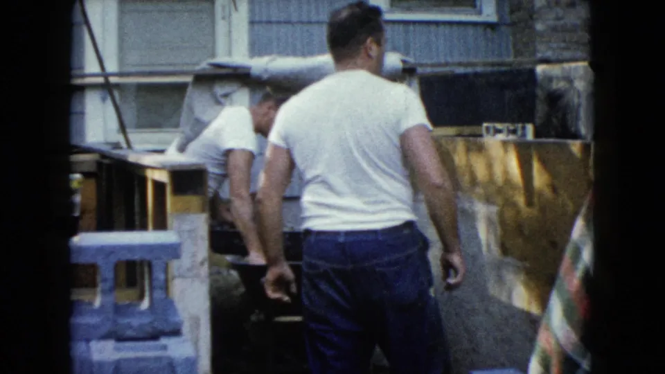 Two adult men continue backyard foundation work, one in the foreground and another bending near cinder blocks and the wheelbarrow.