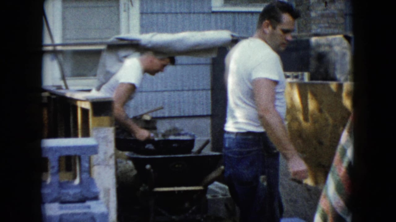 Two men in a backyard near a wheelbarrow and house siding.